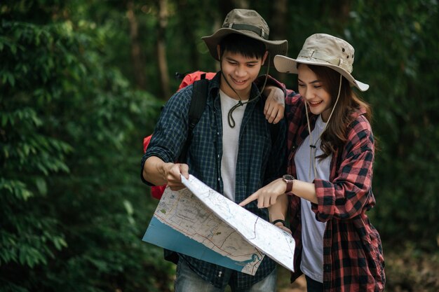 초상 배낭과 트레킹 모자를 쓴 젊은 아시아 미남, 예쁜 여자친구가 숲길을 걷는 동안 종이 지도에 서서 방향을 확인하는 배낭 여행 컨셉입니다.