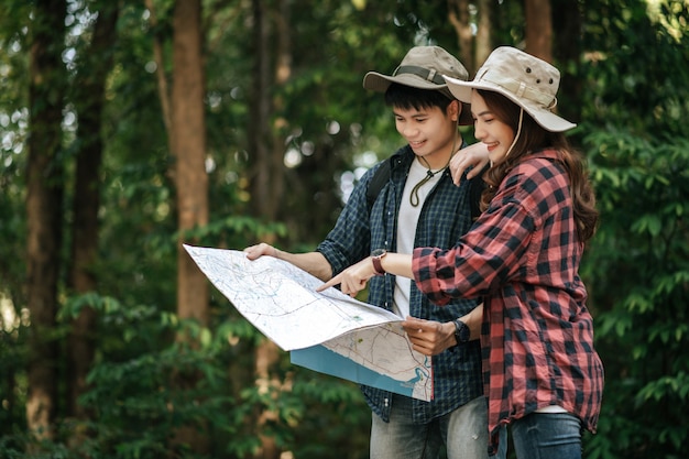 Portrait Young asian handsome man with backpack and trekking hat and pretty girlfriend standing and checking direction on paper map while walking on forest trail, backpack travel concept