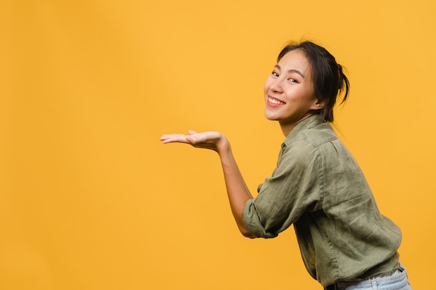 Ritratto di una giovane donna asiatica che sorride con un'espressione allegra, mostra qualcosa di straordinario in uno spazio vuoto in un panno casual isolato su un muro giallo. concetto di espressione facciale.