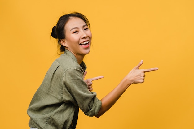 Portrait of young Asian lady smiling with cheerful expression, shows something amazing at blank space in casual cloth  isolated over yellow wall. Facial expression concept.