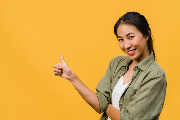 Portrait of young Asian lady smiling with cheerful expression, shows something amazing at blank space in casual cloth  isolated over yellow wall. Facial expression concept.
