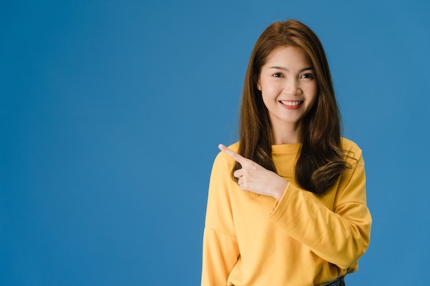 Portrait of young Asian lady smiling with cheerful expression, shows something amazing at blank space in casual clothing and looking at camera isolated over blue background. Facial expression concept.