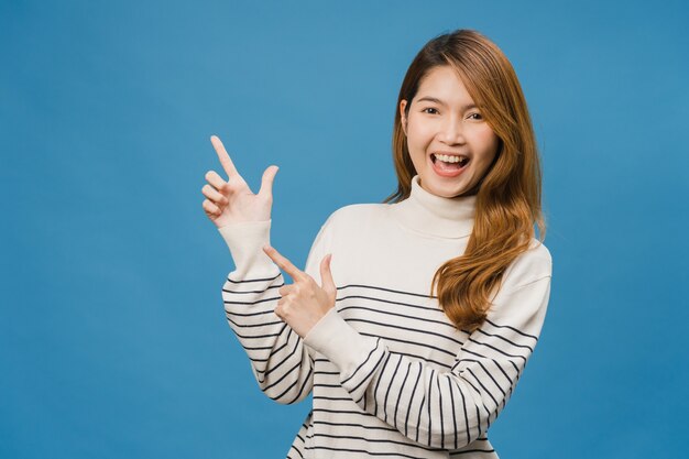 Портрет молодой азиатской леди, улыбающейся с веселым выражением лица, показывает что-то удивительное на пустом месте в повседневной одежде и смотрит на переднюю часть, изолированную над синей стеной