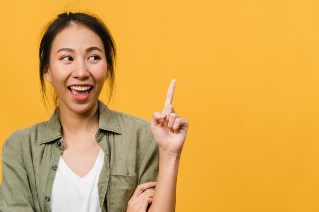 Portrait of young Asian lady smiling with cheerful expression, shows something amazing at blank space in casual clothing and standing isolated over yellow wall. Facial expression concept.