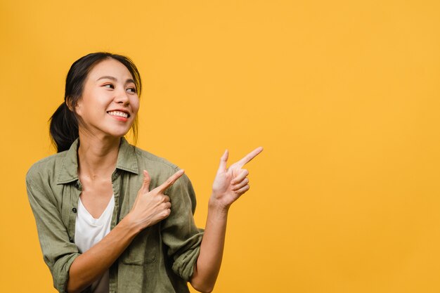 Portrait of young Asian lady smiling with cheerful expression, shows something amazing at blank space in casual clothing and standing isolated over yellow wall. Facial expression concept.