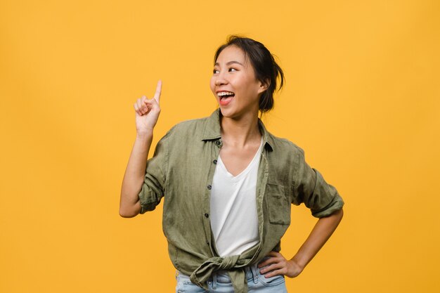 Portrait of young Asian lady smiling with cheerful expression, shows something amazing at blank space in casual clothing and standing isolated over yellow wall. Facial expression concept.