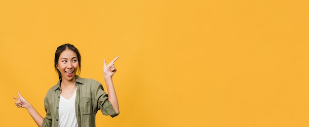 Portrait of young Asian lady smiling with cheerful expression, shows something amazing at blank space in casual clothing and standing isolated over yellow wall. Panoramic banner with copy space.