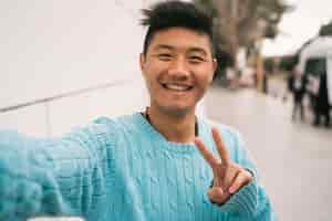 Free photo portrait of young asian man looking confident and taking a selfie while standing outdoors in the street.