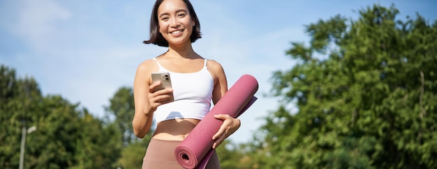 Free Photo portrait of young asian woman checking her phone during workout walking in park with rubber mat