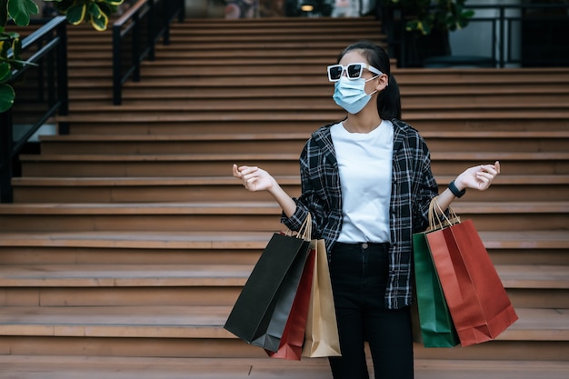 Portrait of young Asian woman in face mask wearing eyeglasses standing on stairs with shopping paper bag