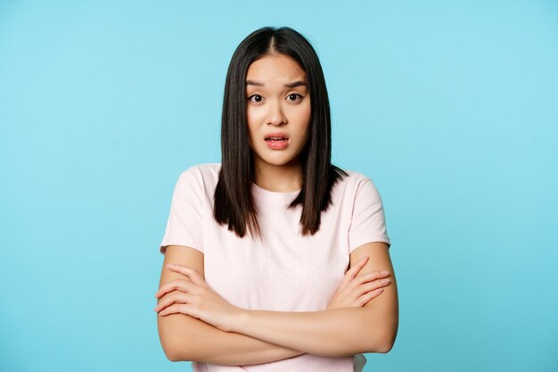 Портрет молодой азиатской женщины, слушающей с запутанным потрясенным выражением лица, стоящей в футболке над ...