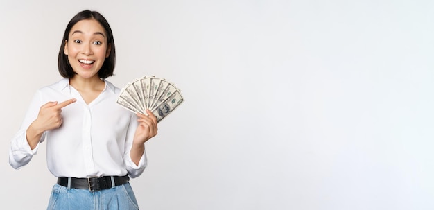 白い背景の上に立っている現金を示す彼女のお金のドルを指している若いアジアの女性の肖像画