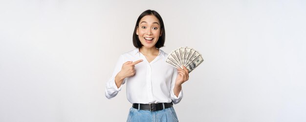 Portrait of young asian woman pointing at her money dollars showing cash standing over white background