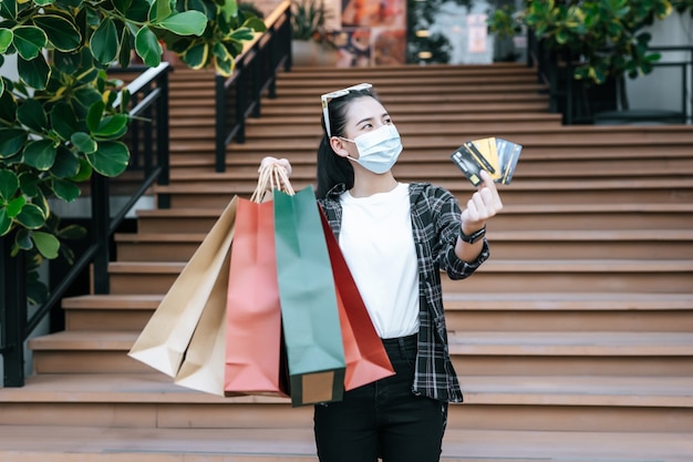 Portrait of young Asian woman in protection mask, eyeglasses on head standing with shopping paper bag