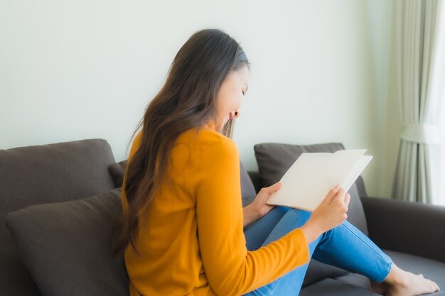肖像若いアジアの女性は、リビングルームで枕とソファ椅子の本を読む