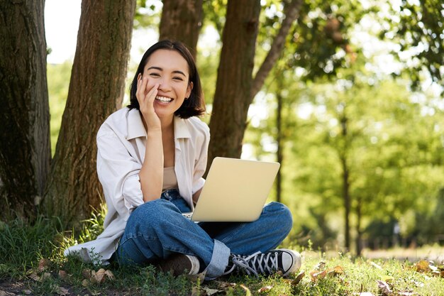 Portrait of young asian woman sitting in park near tree working on laptop using computer outdoors