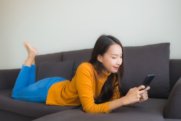 リビングルームの枕が付いているソファーでスマートな携帯電話を使用して肖像若いアジア女性