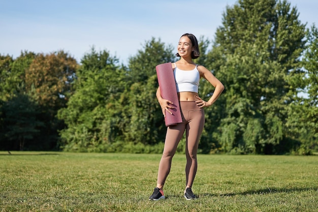 Portrait of young asian woman with rubber mat standing on lawn and looking confident fitness