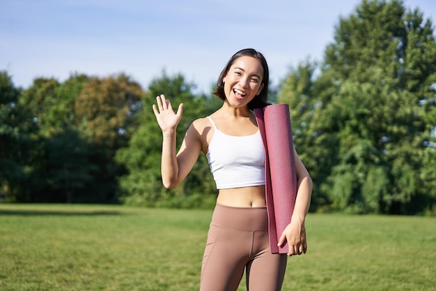 Portrait of young asian woman workout in park outdoors fitness instructor waves hand and smiles invi