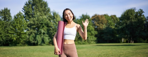 Free photo portrait of young asian woman workout in park outdoors fitness instructor waves hand and smiles