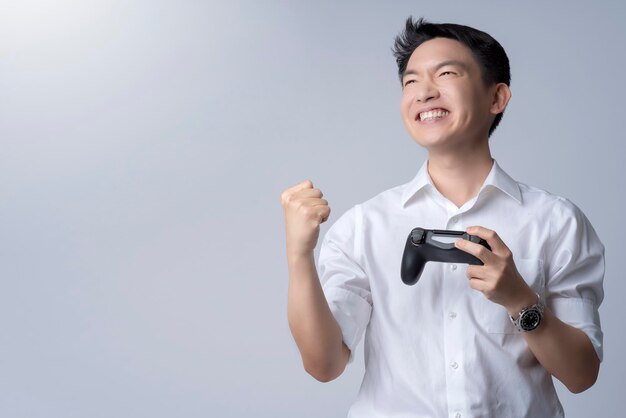 회색 배경에 성공의 승리로 큰 미소를 지으며 조이스틱에서 비디오 콘솔 게임을 하는 젊은 매력적인 아시아 남자의 초상화