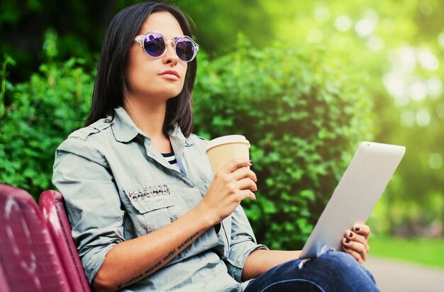 Portrait of young attractive brunette female in sunglasses holds tablet PC drinks coffee in a green summer park.