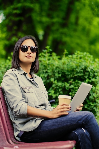 Portrait of young attractive brunette female in sunglasses holds tablet PC drinks coffee in a green summer park.