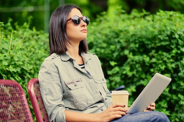 Portrait of young attractive brunette female in sunglasses holds tablet PC drinks coffee in a green summer park.