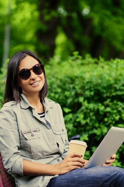 Портрет молодой привлекательной брюнетки в солнцезащитных очках держит планшетный ПК и пьет кофе в зеленом летнем парке.