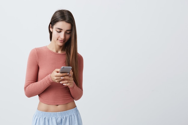 Portrait of young attractive dark-haired caucasian girl with long hair in pink crop top and blue jeans chatting with boyfriend about night date on her smartphone with happy expression.