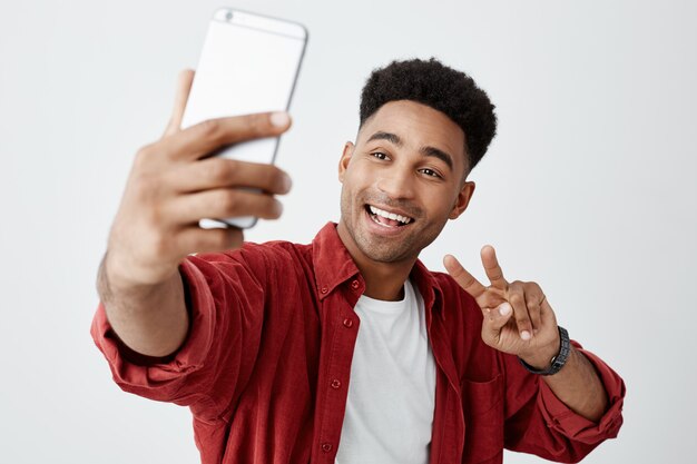 Portrait of young attractive dark-skinned african guy with curly hair in white t-shirt and stylish red shirt talking with girlfriend through video call on cellphone.