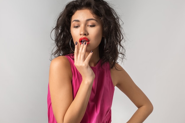 Portrait of young attractive exotic tanned woman in pink stylish dress, red lipstick, sexy, seductive, isolated, summer season style, fashion trend, holding hand at her lips, curly hair