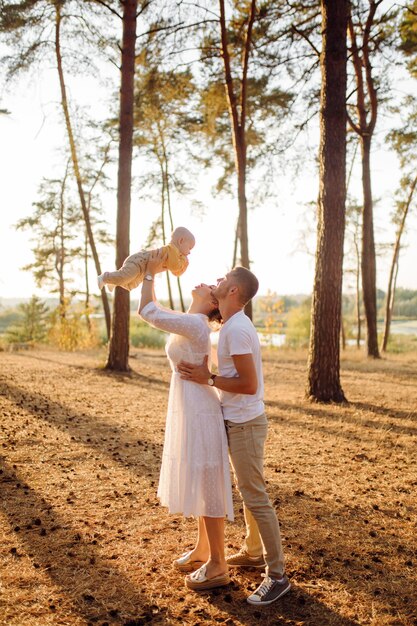 Portrait of young attractive family with little baby son, posing in beautiful autumn pine forest at sunny day. Handsome man and  his pretty brunette wife