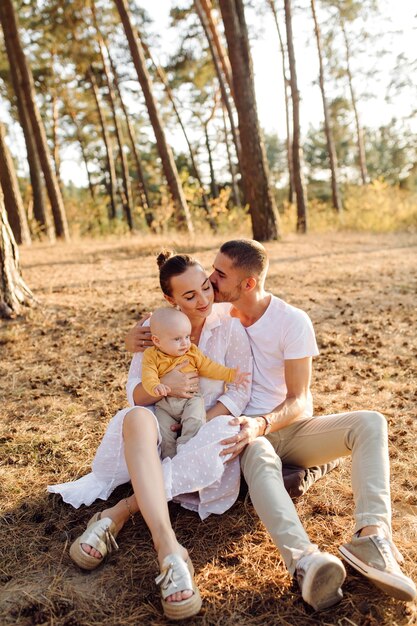 Portrait of young attractive family with little baby son, posing in beautiful autumn pine forest at sunny day. Handsome man and  his pretty brunette wife