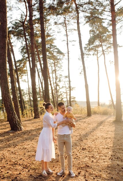 Portrait of young attractive family with little baby son, posing in beautiful autumn pine forest at sunny day. Handsome man and  his pretty brunette wife