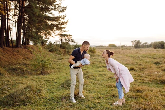 Portrait of young attractive family with little baby son, posing in beautiful autumn pine forest at sunny day. Handsome man and  his pretty brunette wife