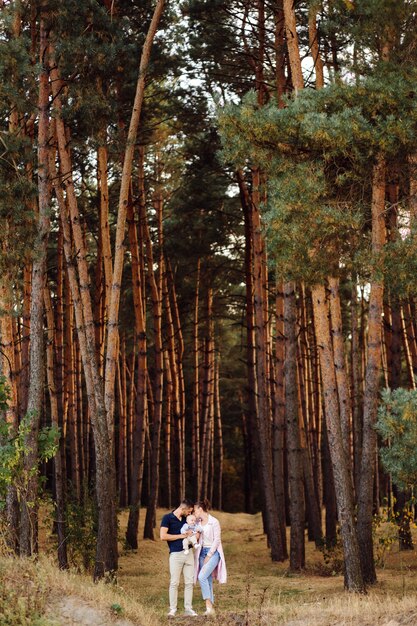 Portrait of young attractive family with little baby son, posing in beautiful autumn pine forest at sunny day. Handsome man and  his pretty brunette wife