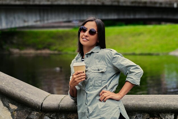 若い魅力的な女性の肖像画は、湖の近くの夏の公園で外出先でコーヒーを飲みます。