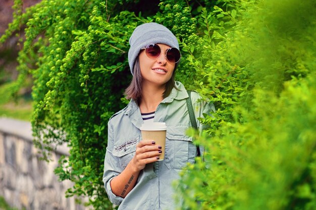 Portrait of young attractive female in sunglasses holds paper coffee cup in a green summer park.