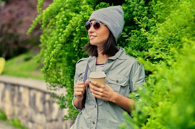 Portrait of young attractive female in sunglasses holds paper coffee cup in a green summer park.