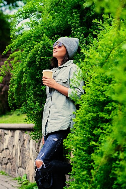 Portrait of young attractive female in sunglasses holds paper coffee cup in a green summer park.