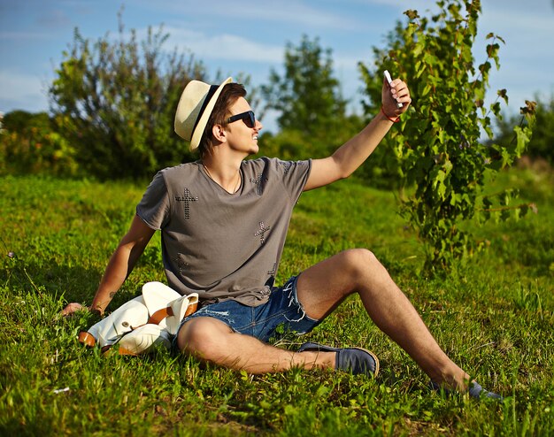 portrait of young attractive modern stylish man in casual cloth in hat in glasses sitting in the park in green grass making selfie