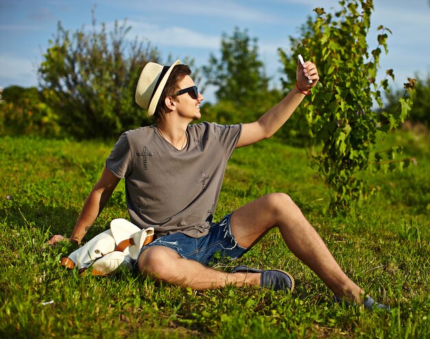 Selfieを作る緑の芝生の公園で座っているメガネの帽子でカジュアルな布で若い魅力的なモダンなスタイリッシュな男の肖像