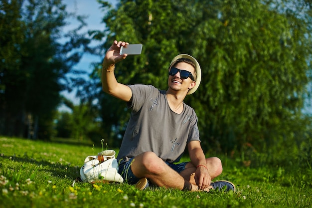 Портрет молодого привлекательного современного стильного человека в повседневной одежде в шляпе в очках, сидя в парке в зеленой траве, делая Селф