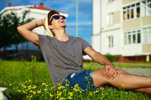 portrait of young attractive modern stylish man in casual cloth in hat in glasses sitting in the park in green grass