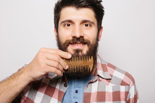 Portrait of young attractive redhead hipster male with serious and confident look, holding wooden comb and doing his thick beard. Stylish bearded barber in checkered shirt combing in salon. Horizontal