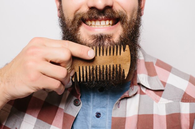 Portrait of young attractive redhead hipster male with serious and confident look, holding wooden comb and doing his thick beard. Stylish bearded barber in checkered shirt combing in salon. Horizontal