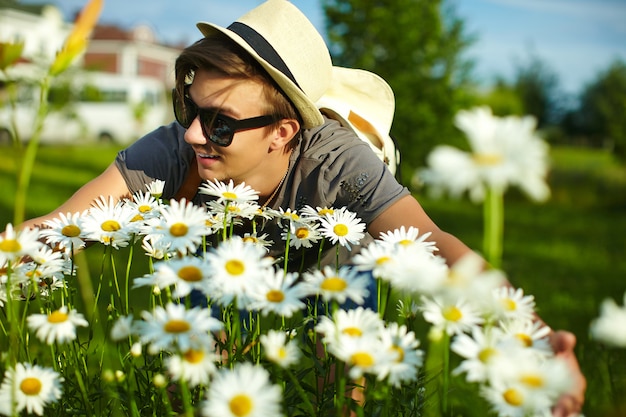 カモミールの明るい色とりどりの花で公園のメガネで帽子のカジュアルな布で若い魅力的な笑みを浮かべてモダンなスタイリッシュな男の肖像