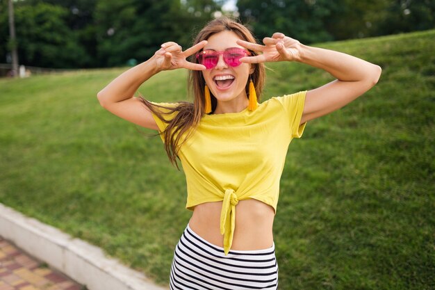 Портрет молодой привлекательной стильной женщины, позирующей в городском парке, улыбающегося веселого настроения, позитива, в желтом топе, полосатой мини-юбке, сумочке, розовых солнцезащитных очках, тенденции моды в летнем стиле
