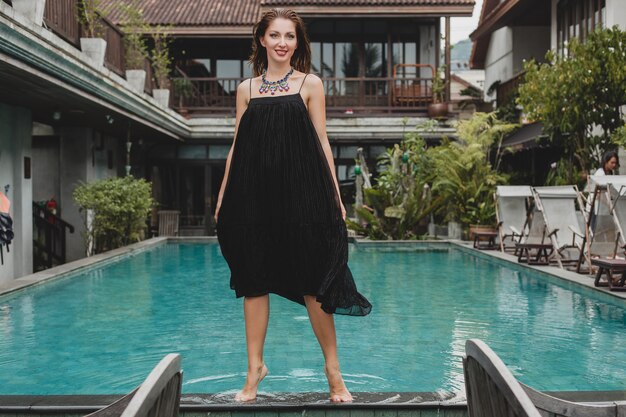 Portrait of young attractive woman in elegant dress, straw hat, summer style, fashion trend, vacation, smiling, stylish accessories, sunglasses, posing on tropical villa on Bali